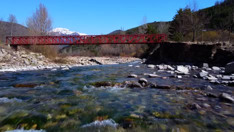 Flying-up-the-river-and-going-under-the-red-bridge-in-the-Spanish-Pyrenees,-with-the-snowy-mountains-in-the-background