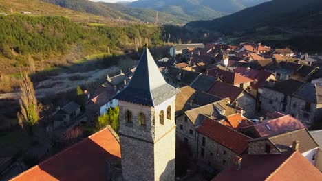 Aerial-video-going-backwards-from-the-church-and-bell-tower-of-the-mountain-village-in-Aragües-del-Puerto,-surrounded-by-green-mountains