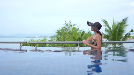 While-leaning-on-the-rail-on-the-edge-of-an-infinity-pool,-a-young-woman-in-a-floppy-sun-hat-and-bathing-suit-looks-from-the-ocean-in-the-background-to-a-point-on-her-left