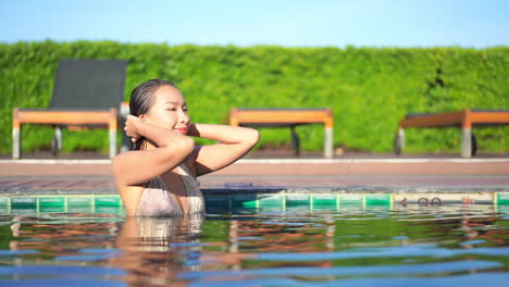 A-passionate-young-Asian-woman-touches-her-wet-hair-inside-the-swimming-pool-water-of-an-exotic-hotel-with-closed-eyes-on-sunset,-portrait-shot