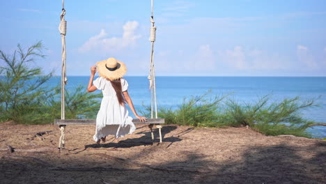 Sitting-on-a-huge-wooden-swing,-a-young-woman-with-her-back-to-the-camera-swings-back-and-forth-as-she-looks-at-the-ocean
