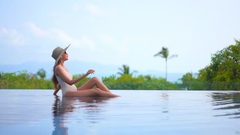 Asian-woman-in-swimming-suit-and-hat-sitting-at-the-edge-of-infinity-pool-leaning-on-one-arm-and-enjoying-the-view-of-tropical-mountains-and-green-trees-in-background,-daytime-slow-motion-side-view