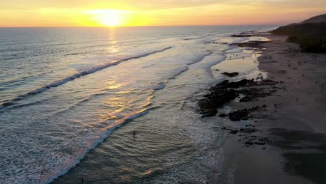 Aerial-flying-across-sunset-reflecting-in-the-water-with-waves-and-surfers-in-the-pacific-ocean-in-Tamarindo,-Costa-Rica