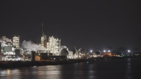 Fábrica-Industrial-Iluminada-Junto-Al-Río-Con-Humo-De-Chimenea-Por-La-Noche