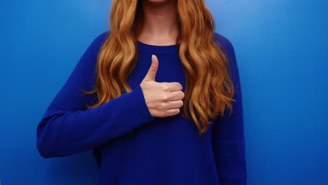 Young-redhead-woman-in-blue-shirt-showing-thumbs-up-gesture,-isolated-on-the-blue-background