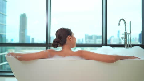 A-young-woman-in-a-spa-like-bathtub-looks-out-the-window-at-a-modern-urban-skyline