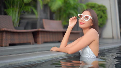 Hermosa-Mujer-Tailandesa-Dentro-Del-Agua-De-La-Piscina-Apoyada-En-El-Lado-De-La-Piscina-Y-Tocando-Sus-Gafas-De-Sol-En-El-Hotel-Resort,-Cámara-Lenta-Estática
