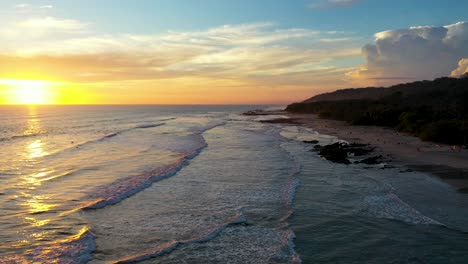 Aerial-flying-from-beach-to-sunset-reflecting-in-the-water-with-waves-and-surfers-in-the-pacific-ocean-in-Tamarindo,-Costa-Rica