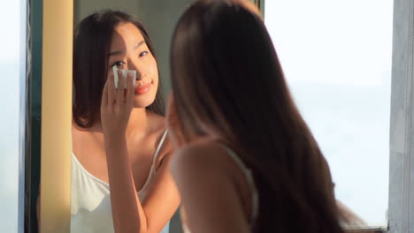 Asian-woman-using-cotton-pads-for-cleansing-the-face-of-cosmetics-with-tonic-or-micellar-cleansing-water-standing-near-the-mirror