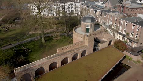 Sunny-day-slow-aerial-pan-showing-top-of-the-historic-Bourgonje-stronghold-tower-and-city-wall-with-park-landscape-in-Zutphen,-The-Netherlands
