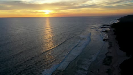 Aerial-of-beautiful-sunset-reflecting-in-the-water-with-waves-and-surfers-in-the-pacific-ocean-in-Tamarindo,-Costa-Rica