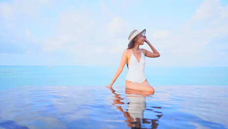 Asian-woman-in-white-monokini-and-skimmer-sun-hat-sitting-on-the-edge-of-the-rooftop-infinity-pool-touching-the-tip-of-the-hat-and-changing-sight,-slow-motion-front-view,-copy-space-template