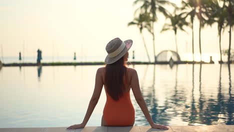 Back-view-of-Fit-female-model-wearing-an-orange-monokini-swimwear-suit-and-hat-sits-on-the-edge-of-the-swimming-pool-leaning-on-arms-at-sunset