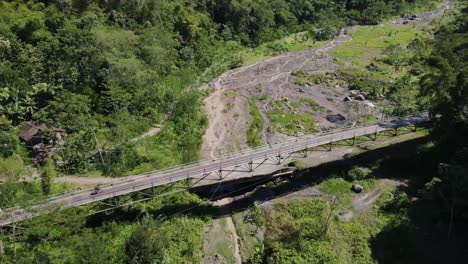 Aerial-view,-cyclists-cross-a-suspension-bridge-and-show-a-panoramic-view-of-Mount-Merapi-when-it-emits-eruption-smoke