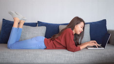 Smiling-Asian-woman-lying-on-sofa-and-typing-on-laptop-keyboard