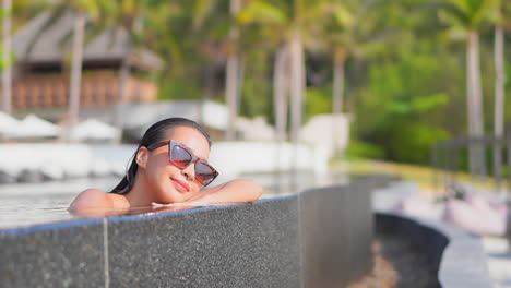Young-Asian-woman-inside-infinity-pool-water-relaxing-leaning-head-on-hands-at-the-edge-of-the-pool,-Face-close-up-slow-motion