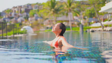 Asian-woman-in-red-bikini-turns-around-with-spread-on-sides-hands-inside-swimming-pool-at-a-private-villa-in-Greece,-slow-motion-daytime-sunny-closed-eyes