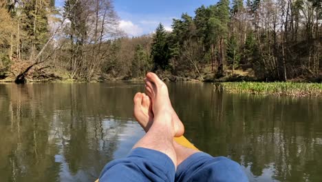 Legs-of-a-man-on-yellow-kayak-bow-sailing-along-tranquil-dark-blue-river-water-against-the-clear-sky-on-summer-day-closeup