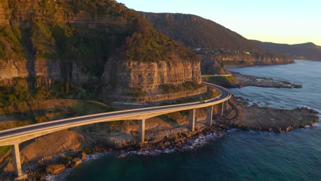 Modern-Highway-Of-Sea-Cliff-Bridge-Around-Steep-Sandstone-Cliffs-On-Australian-Pacific-Coast-During-Sunset---vertical-aerial-panorama