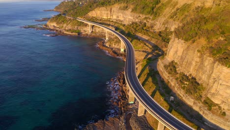 Sea-Cliff-Bridge-Section-Of-The-Grand-Pacific-Drive-Around-Steep-Sandstone-Cliff-At-The-Edge-Of-Pacific-Coast-During-Sunset---aerial-drone-shot