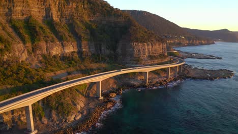 Cars-Driving-By-The-Sea-Cliff-Bridge-In-NSW-Australia-In-Sunrise---aerial-shot