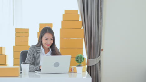 Beautiful-business-Asian-woman-typing-on-laptop-in-office-with-piles-of-cardboard-boxes-in-background