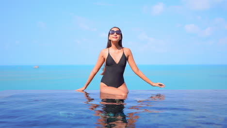Young-Woman-in-Black-Swimming-Suit-Sitting-on-the-Edge-of-Infinity-Pool-and-Touching-the-Frame-of-Sunglasses-on-Turquoise-Seascape-Background-on-a-Sunny-Day-Slow-Motion-Front-View