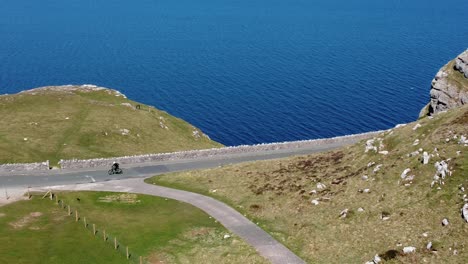 Tourist-riding-mountain-bike-along-scenic-green-mountain-country-road-overlooking-gorgeous-blue-Irish-sea-pan-left