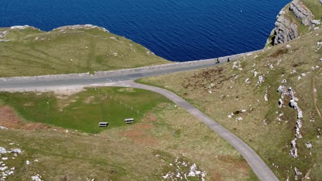 Tourist-riding-mountain-bikes-along-scenic-green-mountain-country-road-overlooking-gorgeous-blue-Irish-sea-aerial-panning-right