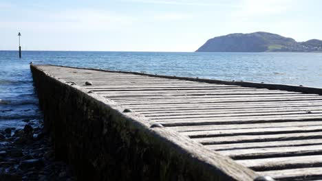 Worn-wooden-boardwalk-leading-down-to-shimmering-North-Wales-island-blue-ocean-waves-rising-right-shot