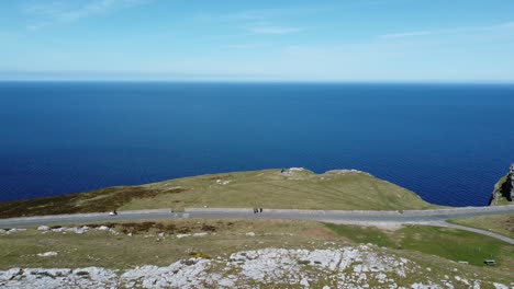 Tourist-hikers-walk-along-scenic-green-mountain-country-road-overlooking-gorgeous-blue-Irish-sea-aerial-panning-right-shot