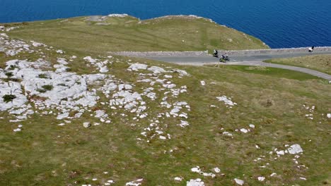 Tourists-riding-mountain-bike-along-scenic-green-mountain-country-road-overlooking-gorgeous-blue-Irish-sea-Aerial-rising-dolly-left