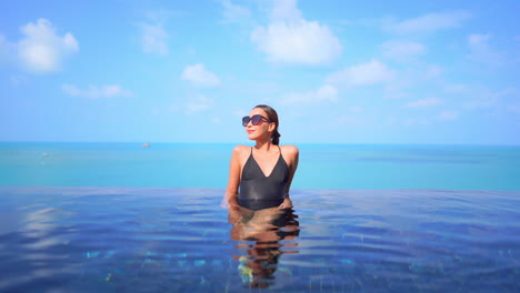 Fit-Woman-Model-in-Black-Swimming-Suit-Leaning-Arms-on-the-Edge-of-Infinity-Pool-on-Turquoise-Seascape-Background-Wearing-Sunglasses-on-a-Sunny-Day-Slow-Motion