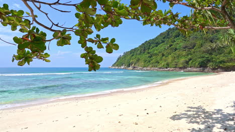 Empty-White-sand-island-beach-with-a-mountain-on-Background-and-tropical-tree-branch-in-the-foreground-on-a-sunny-day-in-Thailand