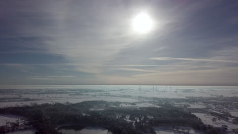 Luftaufnahme-Der-Verschneiten-Winterlandschaft-Mit-Waldbäumen-Und-Windturbinenpark-Im-Hintergrund-Bei-Sonnenlicht