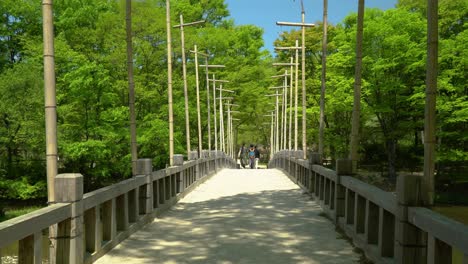 Amigos-Caminando-En-Un-Viejo-Puente-De-Madera-Con-Frondosos-árboles-Verdes-En-El-Pueblo-Folclórico-Coreano-En-Yongin,-Seúl,-Corea-Del-Sur