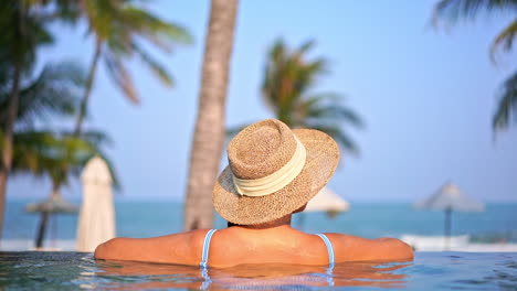 Back-view-of-woman-with-straw-summer-hat-relaxing-in-seafront-infinity-spa-pool