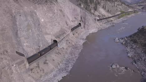 Railroad-train-of-open-hopper-cars-haul-ore-thru-river-canyon-tunnels