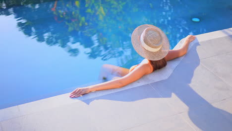 Sexy-woman-sitting-on-edge-of-swimming-pool-of-exotic-hotel-wearing-straw-hat
