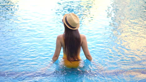 Backside-view-of-an-unrecognizable-woman-standing-inside-swimming-pool-water-wearing-fedora-straw-hat-at-an-exotic-hotel-in-Florida