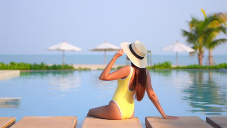 Beautiful-young-woman-in-yellow-swimsuit-with-sunhat-sits-on-edge-of-luxury-swimming-pool