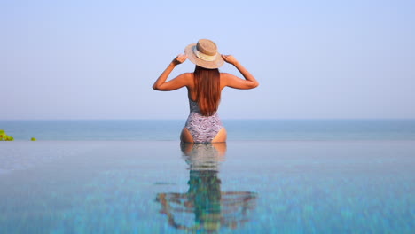 Back-of-a-woman-in-swimwear-sitting-on-infinity-pool-border,-touching-her-sun-hat-and-raising-hands-up-with-a-stunning-view-of-tropical-sea-horizon-in-Miami,-Florida,-static