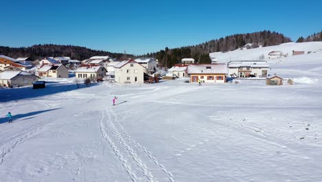 Aerial-tracking-shot-of-biathlon-athlete-in-pink-ski-suit-skiing-uphill-snowy-track-beside-apartment-houses-in-Jura,Lajoux,France