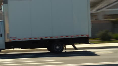 Home-moving-and-shipping-company-white-box-truck-driving-down-a-road-to-its-destination-in-4K---panning-shot