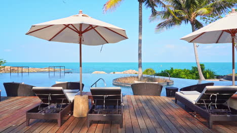 Erstaunliche-Exotische-Infinity-Pool-Lounge-Mit-Kokospalmen-Auf-Der-Felseninsel-Bali,-Liegestühlen-Und-Weißen-Sonnenschirmen-Mit-Kleinen-Tischen-Davor