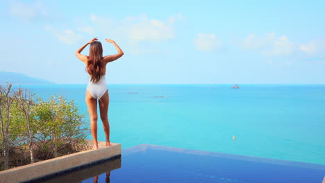 Petite-female-in-white-swimsuit-standing-above-infinity-pool-and-raising-hands,-turquoise-tropical-sea-and-horizon-in-background,-slow-motion