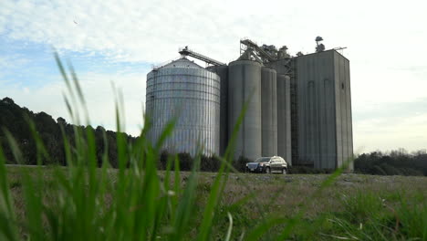 A-grain-silo-towers-over-the-nearby-countryside-in-spring-as-cars-drive-by---sliding,-low-angle-view-with-grass-in-the-foreground