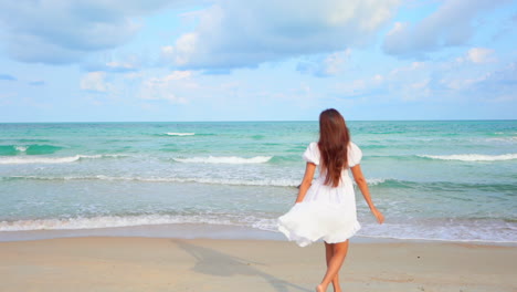 Back-of-attractive-woman-in-fluttering-summer-dress-walking-on-sand-beach-by-sea-looking-at-skyline-and-raising-hands,-slow-motion