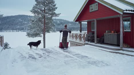 Man-Shoveling-Snow-Off-In-A-Wood-Patio-Of-A-Rustic-Cottage-With-His-Dog-In-Trondheim,-Norway