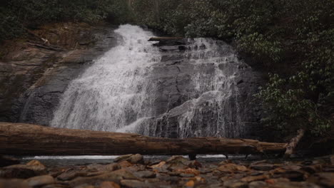 Malerischer-Wasserfall-Im-Wald,-Der-In-Zeitlupe-Einen-Berghang-Hinunterstürzt,-Mit-Einem-Baumstamm-Im-Vordergrund---Gleitender-Blick